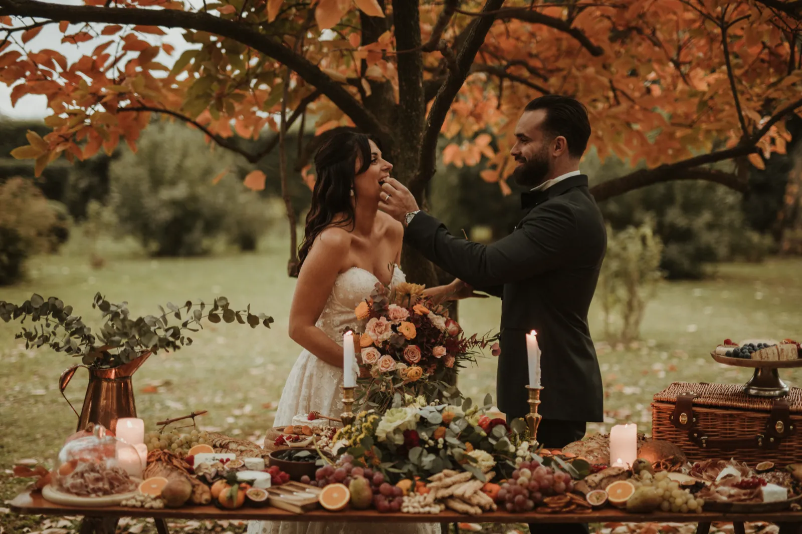 Mariés se partageant une bouchée devant une longue table de pâturage dressée sous un arbre en automne
